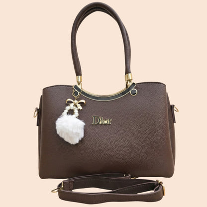 Brown handbag with a white pom-pom charm and gold accents on a beige background