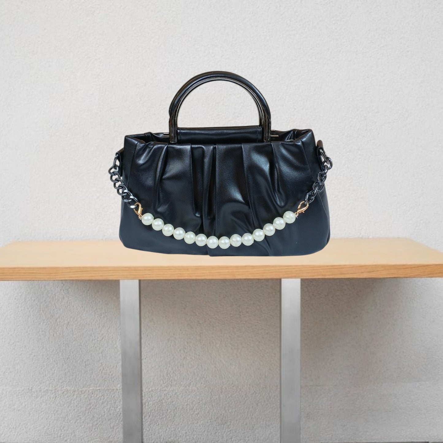 Black handbag with a pearl chain on a wooden table against a light gray wall.