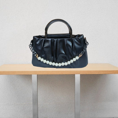 Black handbag with a pearl chain on a wooden table against a light gray wall.